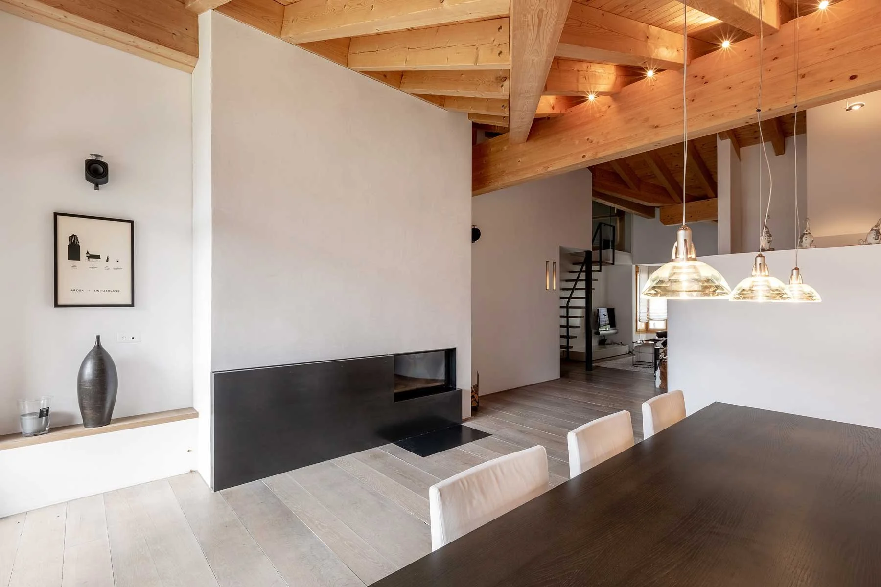 Chalet Innenansicht eines modernen Wohn- und Esszimmers mit Holzdecke, weißen Wänden, einer schwarzen Kaminwand, einem Esstisch mit Stühlen, Hängeleuchten und minimalistischer Dekoration.