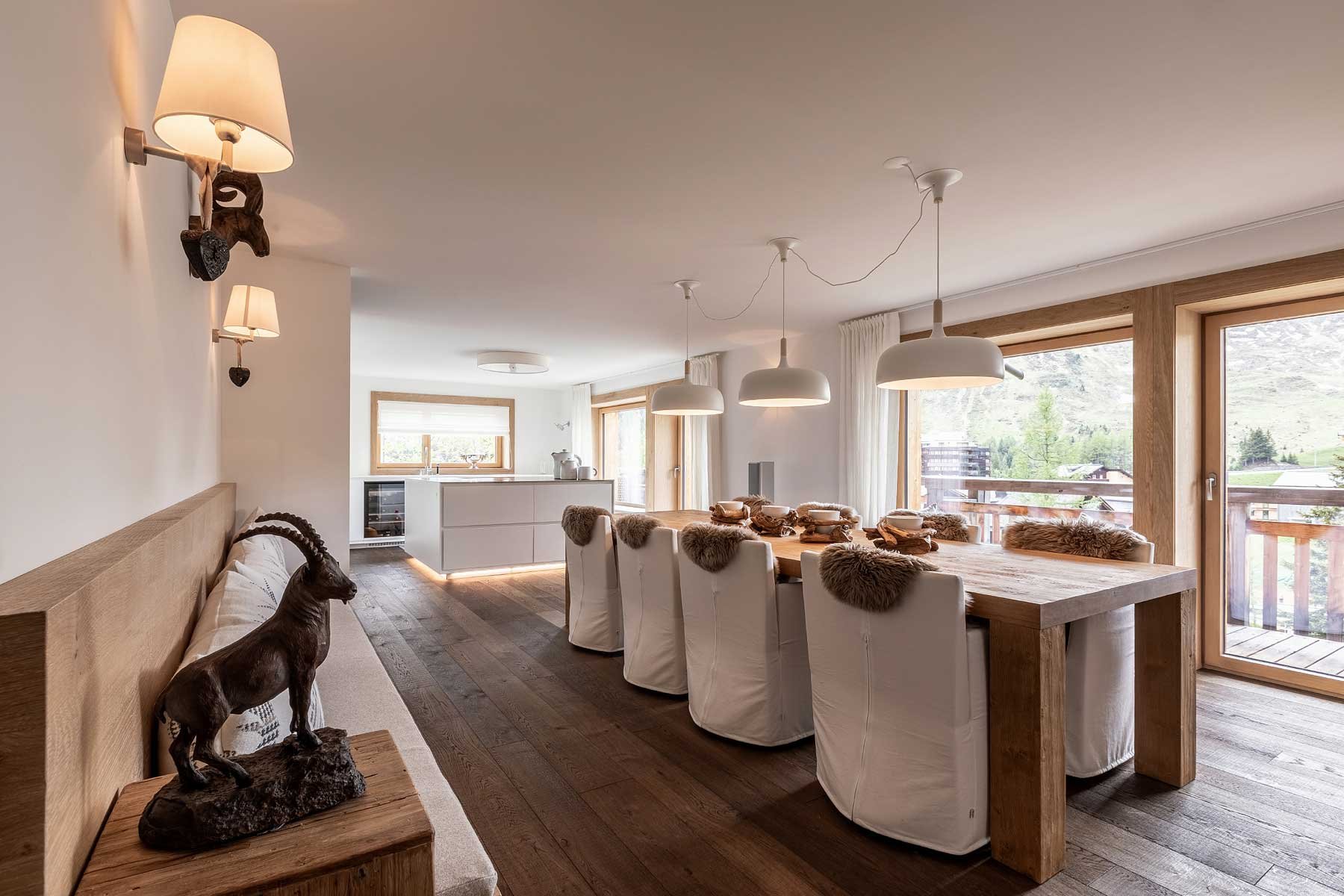 Ein offener, gemütlicher Raum im Chalet-Stil mit Holzdetails, einem großen Esstisch mit weißen Stuhlen und Felldecken, großen Fenstern mit Blick auf die Berge, led-lampen und Wandlampen, einer kleinen Kücheninsel im Hintergrund, dekorativen Skulpturen und Naturmaterialien.
