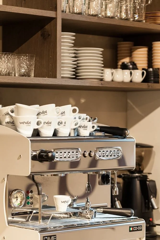 Eine Espressomaschine mit mehreren weißen Kaffeetassen, darunter eine kleine Kaffeetasse, und einem Regal mit Geschirr wie Gläsern, Tellern und Tassen im Hintergrund.