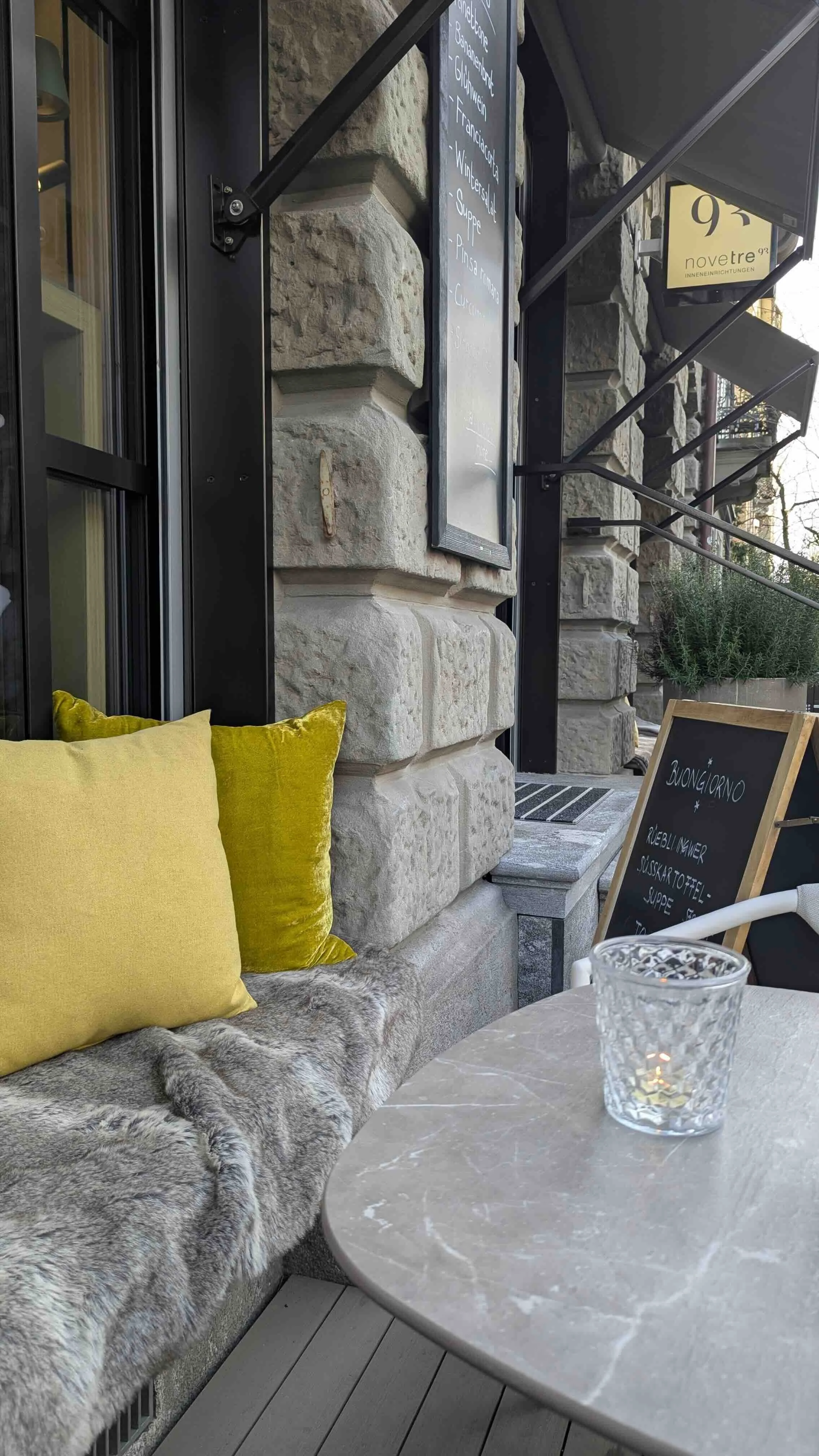 Außensitzbereich eines Cafés mit einer grauen Marmortisch, einem Glas Kerzenhalter, gelben Kissen auf einer Polsterbank, einer schwarzen Tafel mit Tagesangeboten, einer schwarzen Tafel, Steinmauer und Treppe, ein Schild mit der Aufschrift 'novetre Inneninrichtungen'.