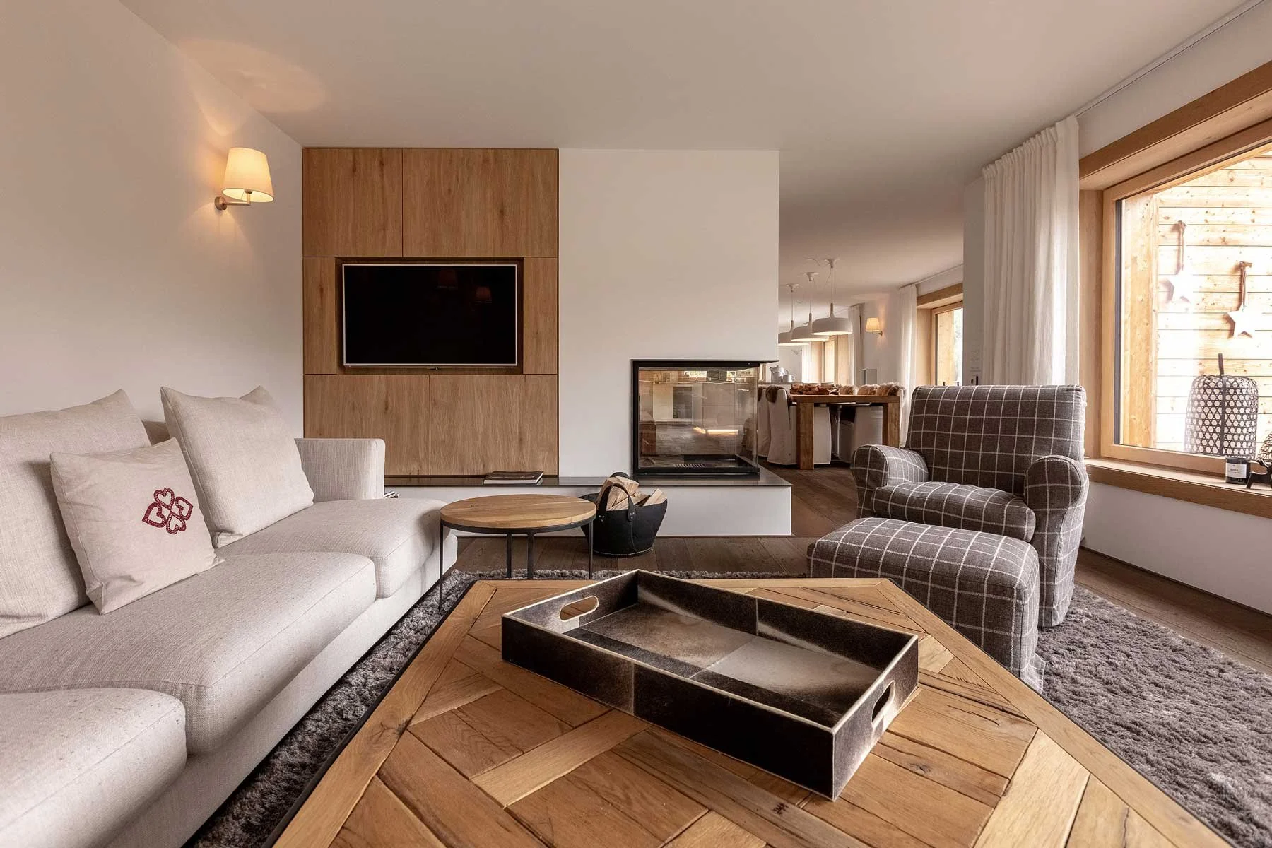 Ein modernes Wohnzimmer mit einem grauen Sofa, Eckkissen und einem Holztisch. Große Fenster lassen viel Licht herein, und es gibt einen Kamin mit Glasabdeckung. Der Boden ist aus Holz, und die Wände sind weiß, mit Wandlampen und Vorhängen. Im Hintergrund ist ein Essbereich sichtbar.