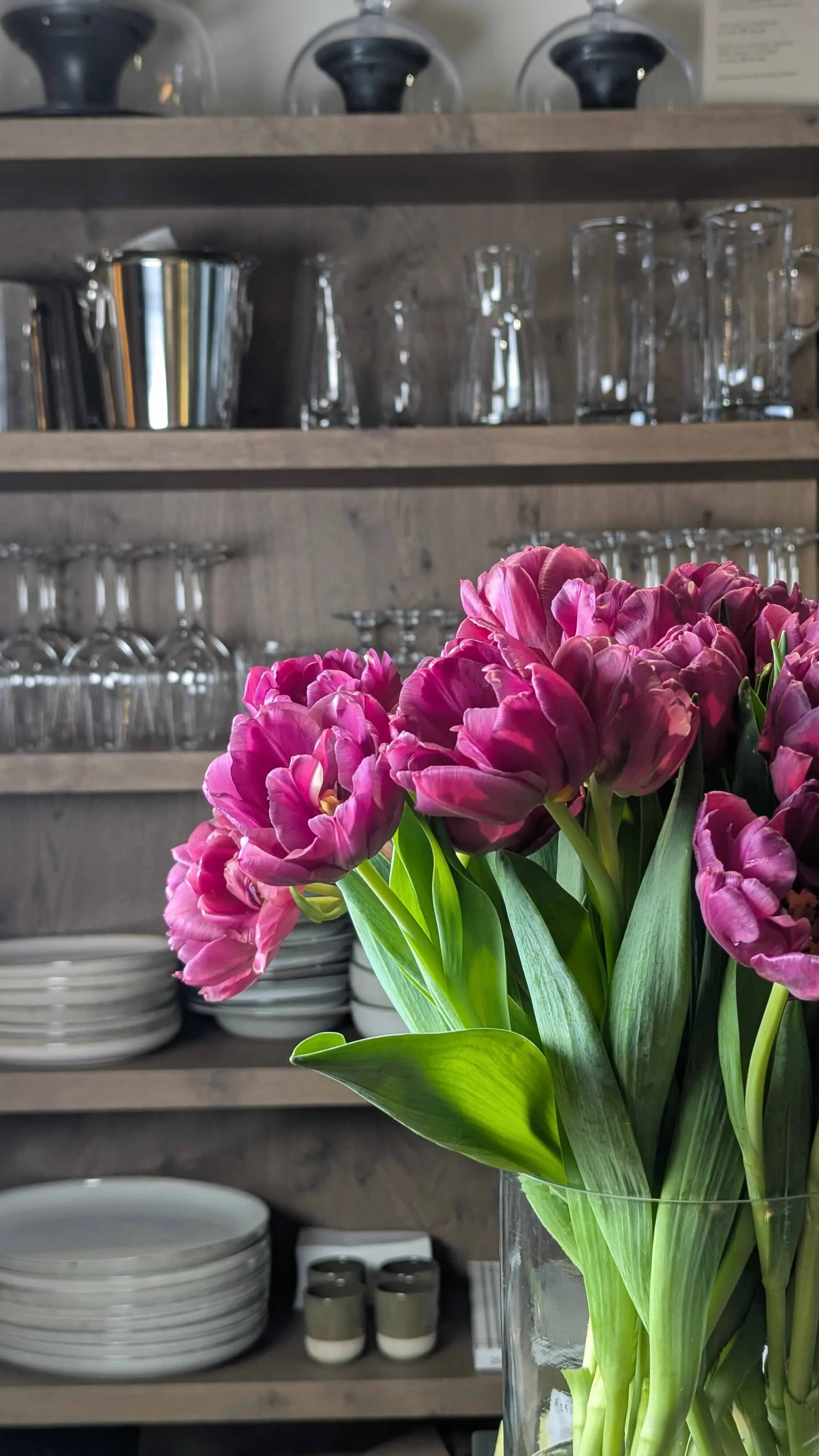 Ein Blumenstrauß aus pinkfarbenen Tulpen in einer Glasvase. Im Hintergrund sind Geschirr und Gläser auf einer Holzregal.