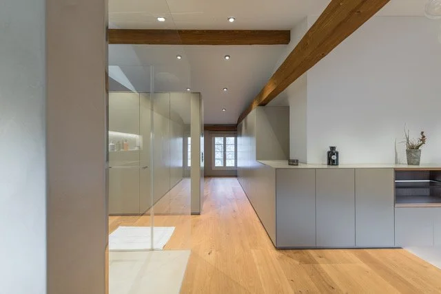 Moderner Chalet Innenraum mit Holzboden, weißen Wänden und sichtbaren Holzbalken an der Decke, Küchen- oder Wohnzimmerbereich mit Stauraum und Fenster