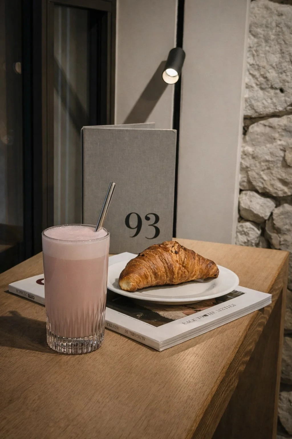 Ein Glas mit rosa Getränk und Strohhalm, ein croissant auf einem weißen Teller, ein graues Buch mit der Nummer 93, auf einem Holztisch in einem Café.
