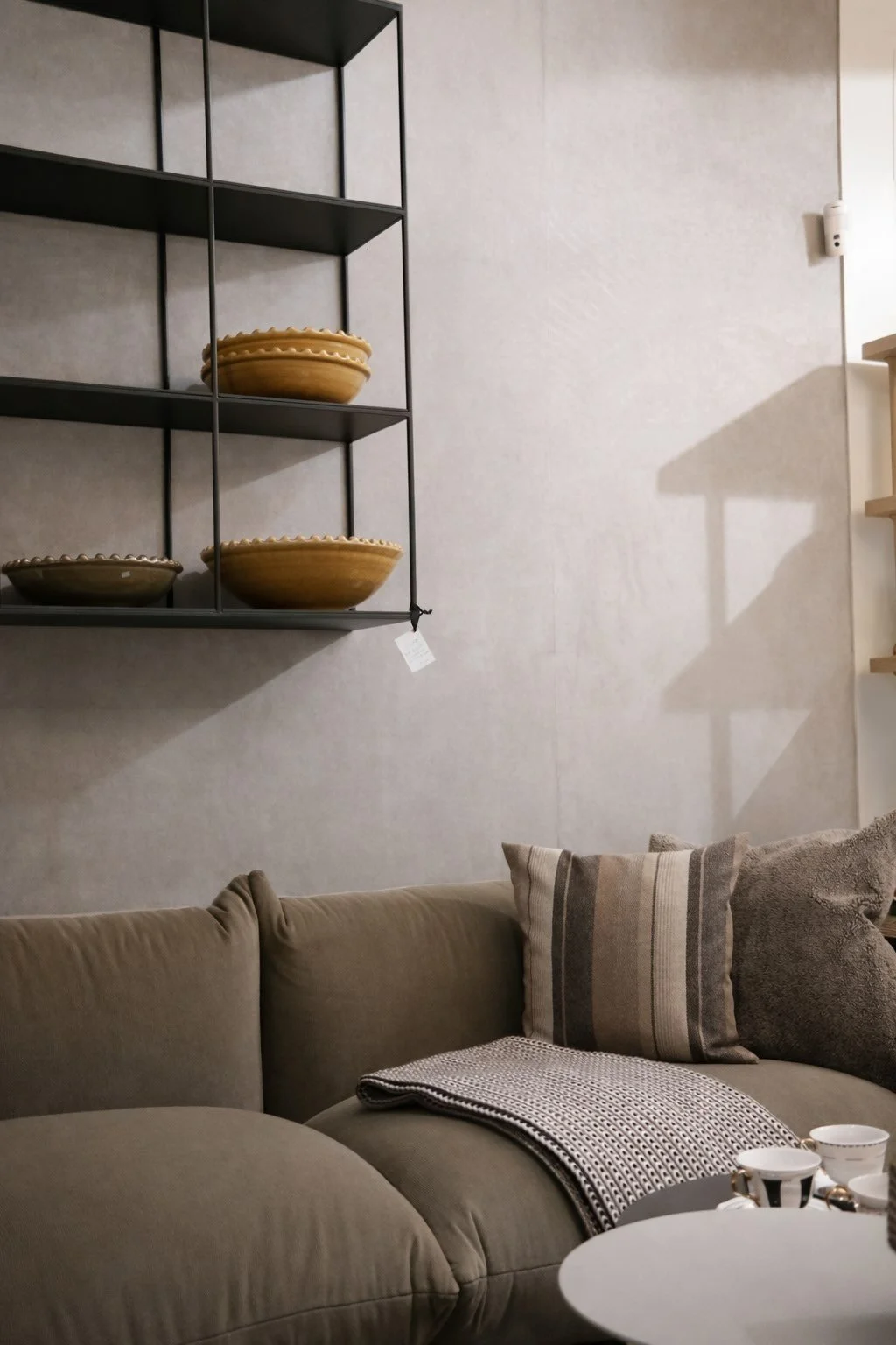 Wohnzimmer mit grauer Couch, Kissen, Decke, einem runden schwarzen Tisch mit Teegeschirr, einer Wandregal mit drei goldfarbenen Schalen, indirekte Beleuchtung, weiße Wand
