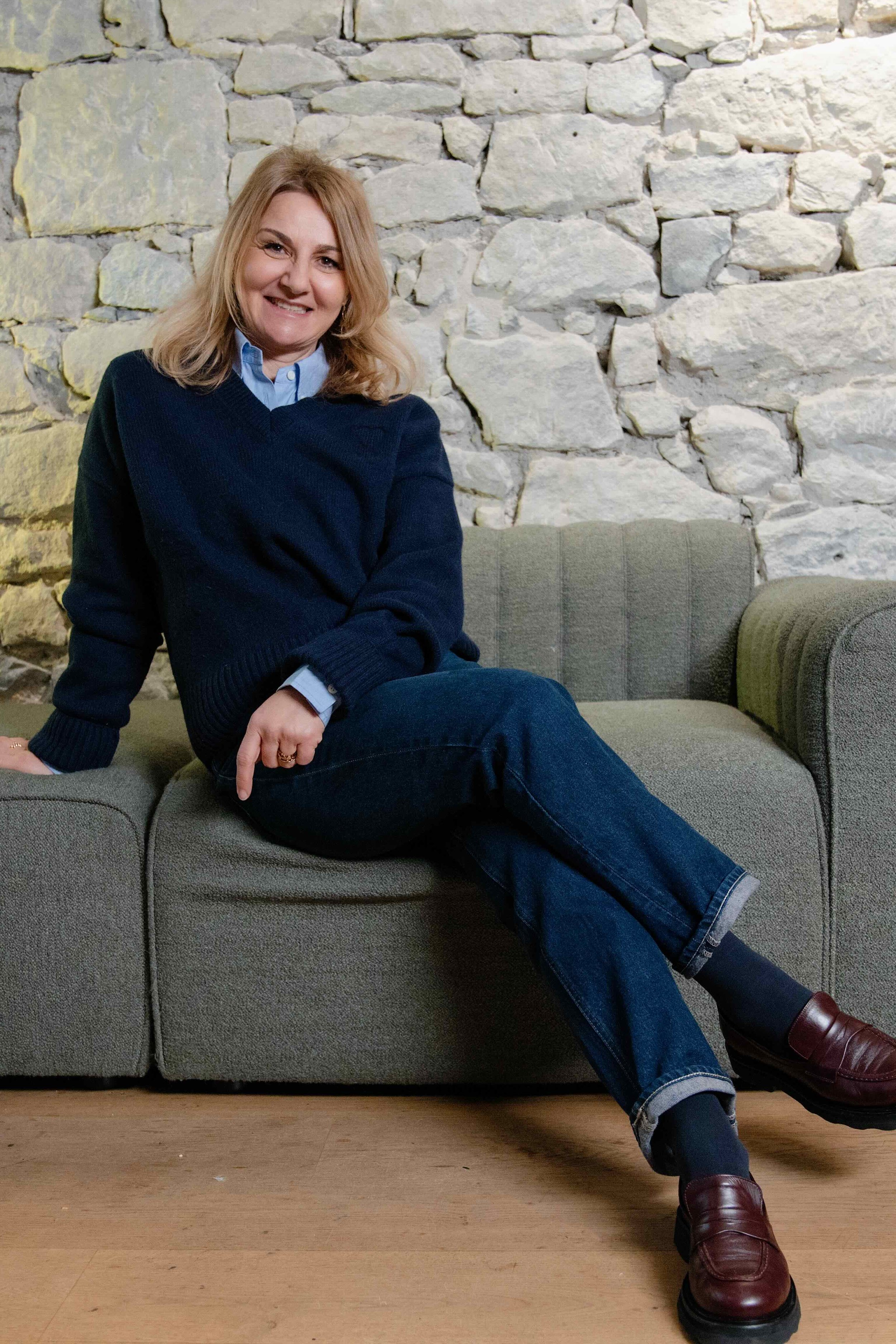 Eine Frau mit blonden Haaren sitzt auf einem grauen Sofa vor einer Steinwand und lächelt in die Kamera.