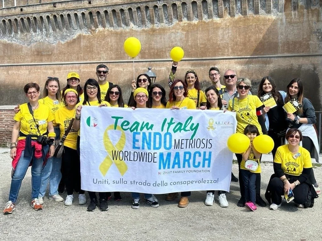 Un gruppo di persone che indossano magliette gialle e tengono un grande striscione durante un evento di sensibilizzazione sulla endometriosi, con alcuni che tengono palloncini gialli, di fronte a un muro di pietra.