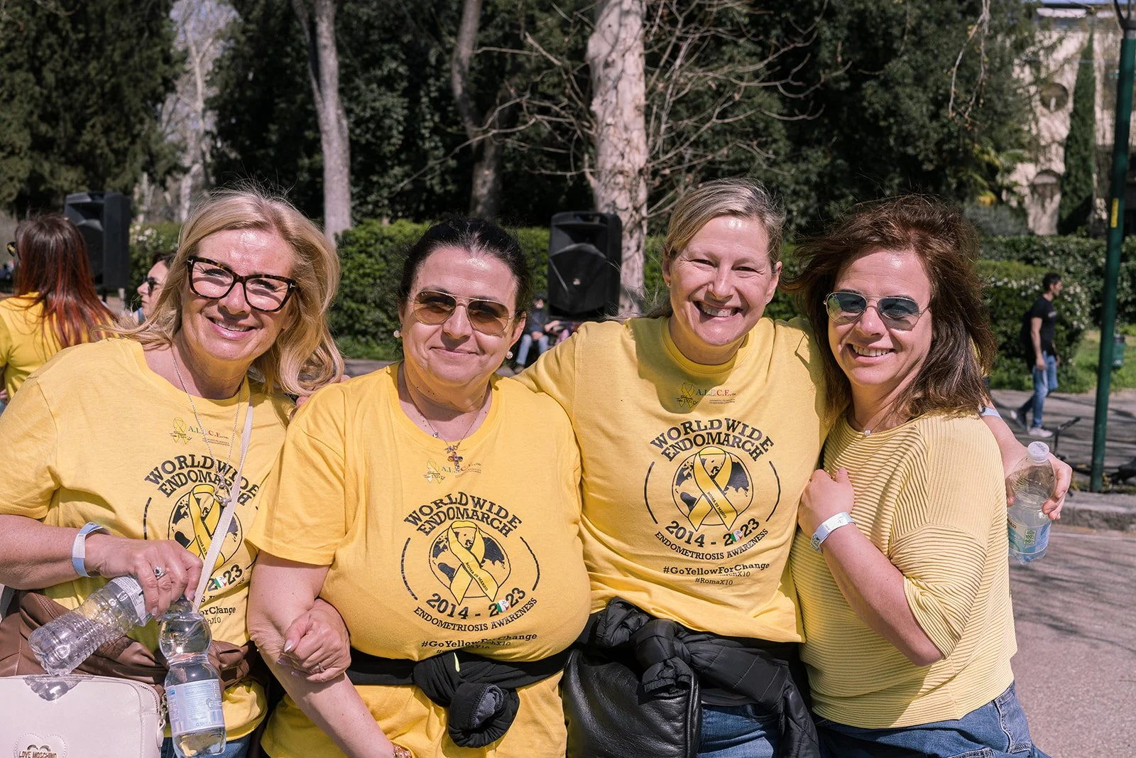 Quattro donne che partecipano a una maratona per la consapevolezza dell'endometriosi, indossano magliette gialle con il logo e le informazioni dell'evento, sorridenti e posando per la foto all'aperto.