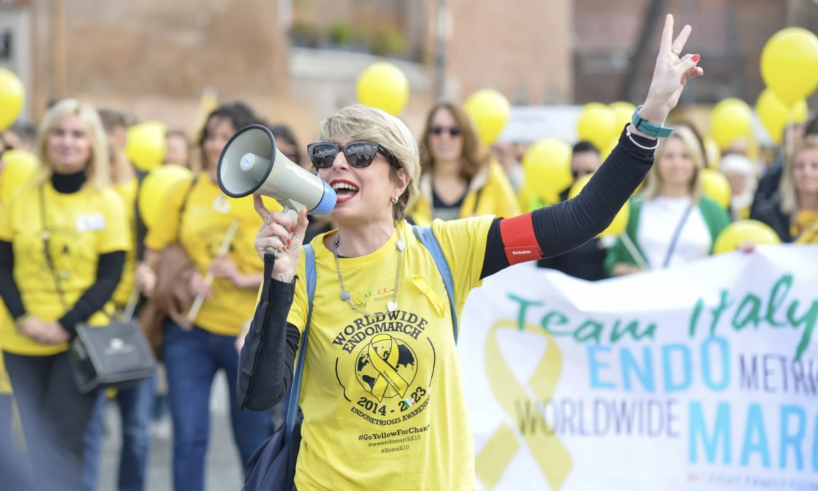 Donna con occhiali da sole parla in un megafono durante una marcia per la consapevolezza dell'Endometriosi, con molte persone che indossano magliette gialle e portano palloncini gialli sullo sfondo.