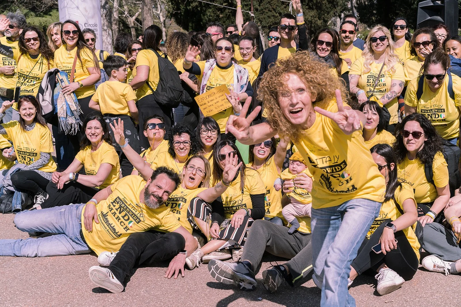 Un gruppo di persone in maglietta gialla che partecipano a una passeggiata o evento sociale, sorridenti e che salutano alla fotocamera in un ambiente all'aperto