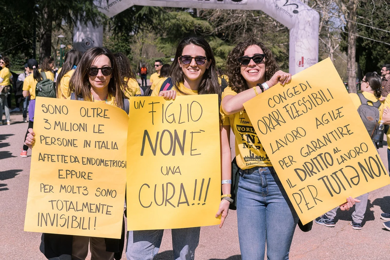 Tre donne alzate in mano cartelli gialli durante una manifestazione per i diritti delle donne, con altre persone sullo sfondo.