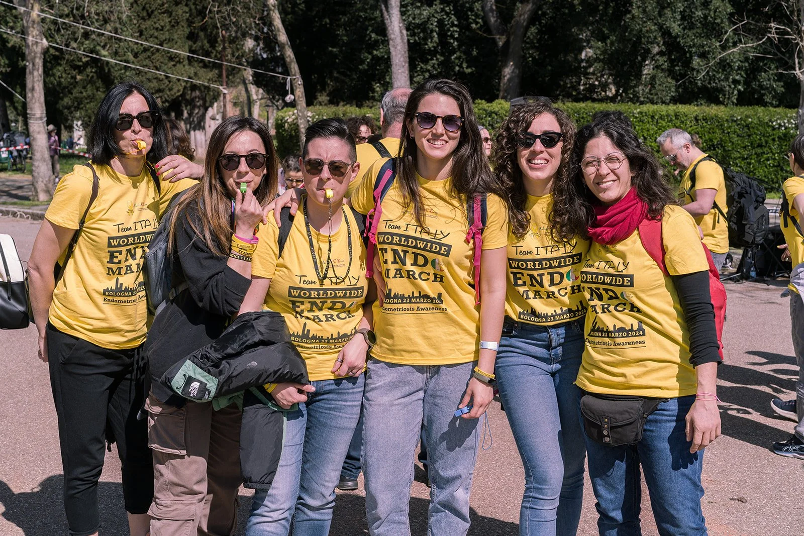 Gruppo di persone che partecipano a un evento di sensibilizzazione sulla endometriosi, indossando magliette gialle con scritte e simboli correlati, all'aperto sotto un cielo soleggiato.