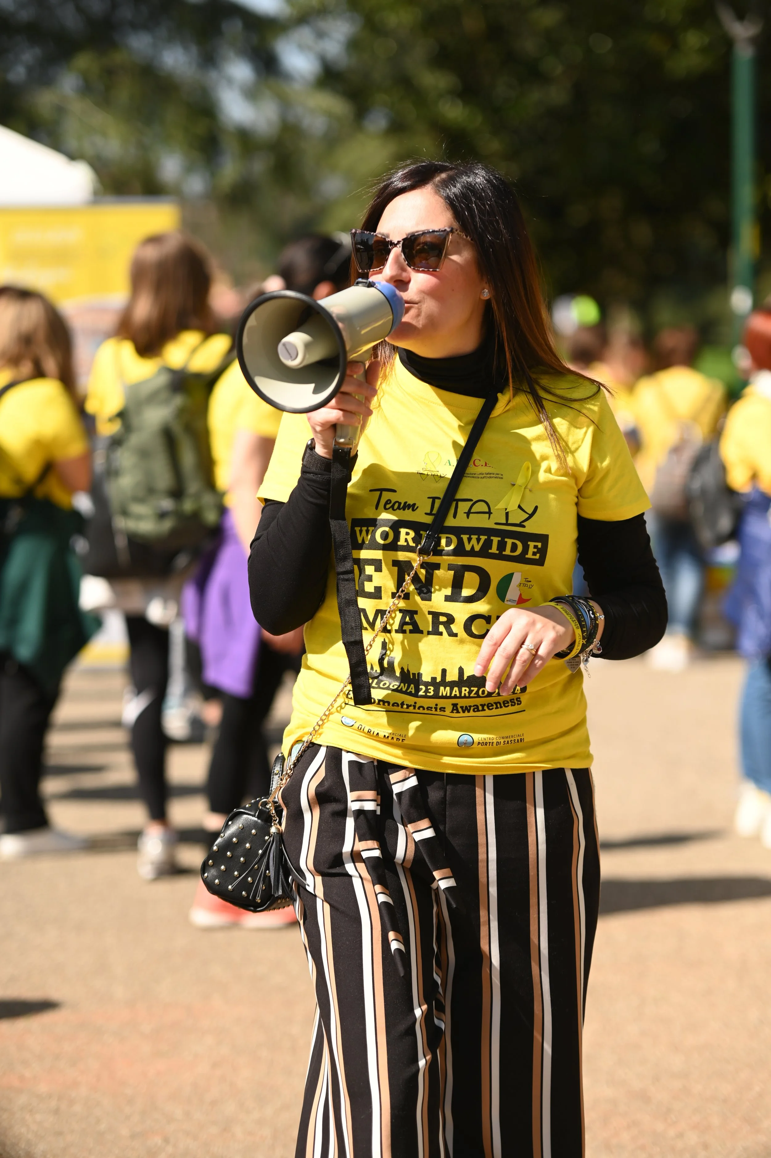 Donna con occhiali da sole che parla in un megafono durante un evento all'aperto con persone vestite di giallo che partecipano.