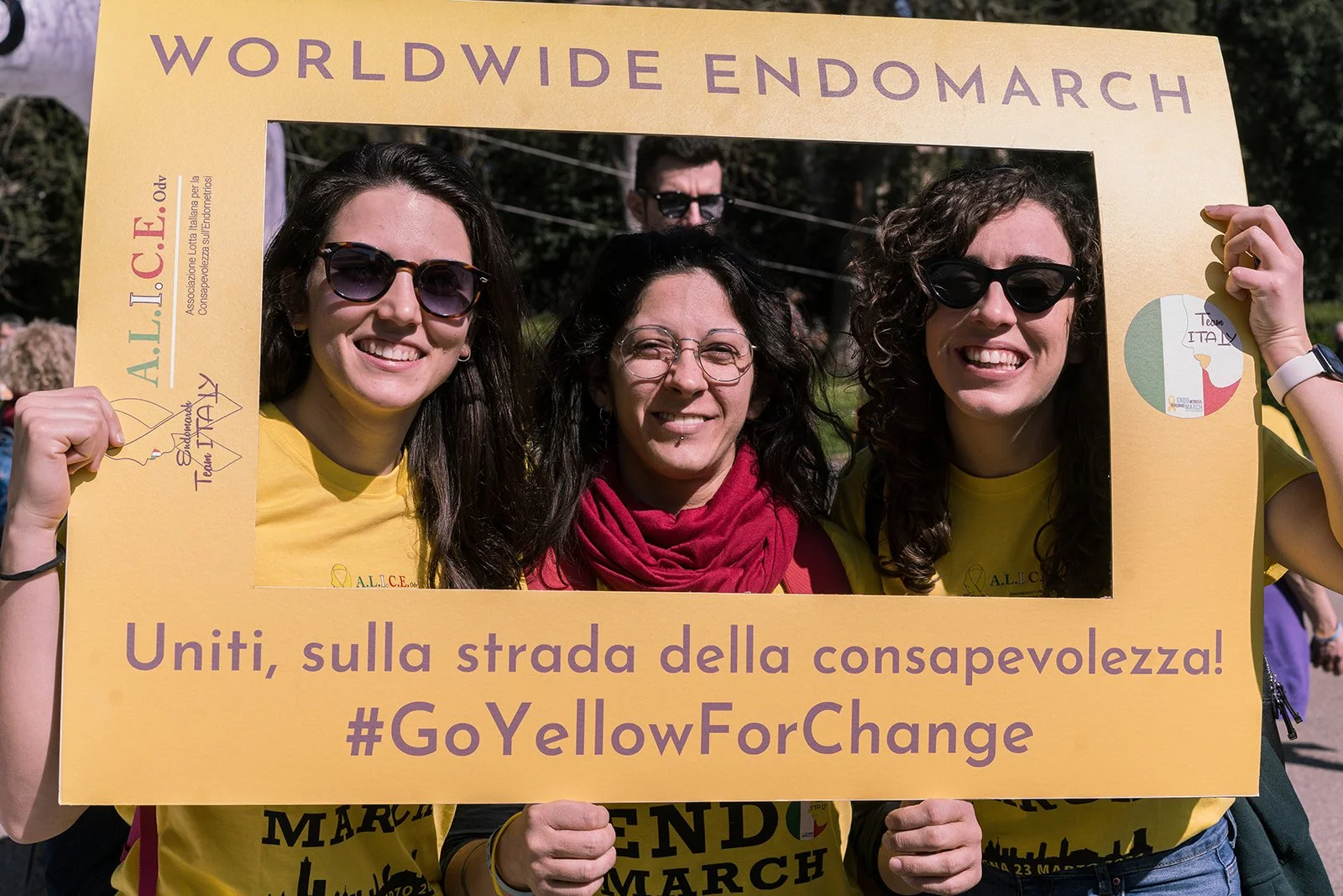 Tre persone sorridenti che tengono un grande telaio giallo con scritto 'WHOEVER ENDOMARCH' e 'Uniti, sulla strada della consapevolezza! #GoYellowForChange' durante una manifestazione.