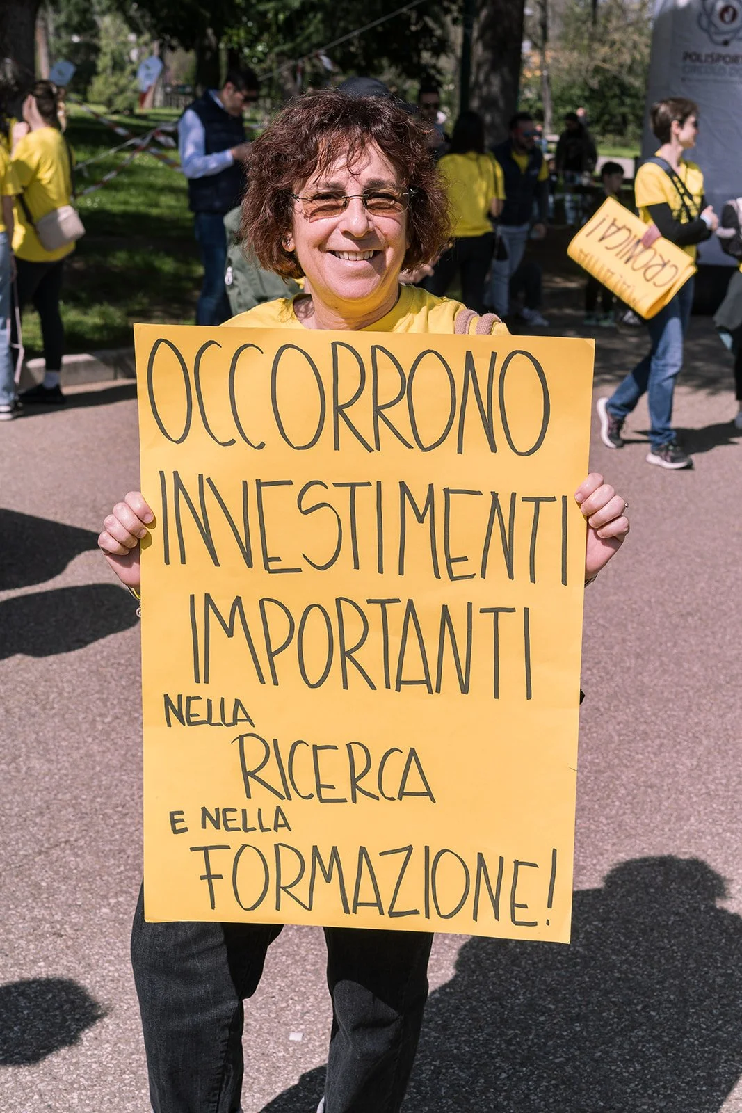 Una donna sorridente tiene un cartello giallo con scritte in italiano durante una manifestazione, con altri partecipanti e alberi sullo sfondo.