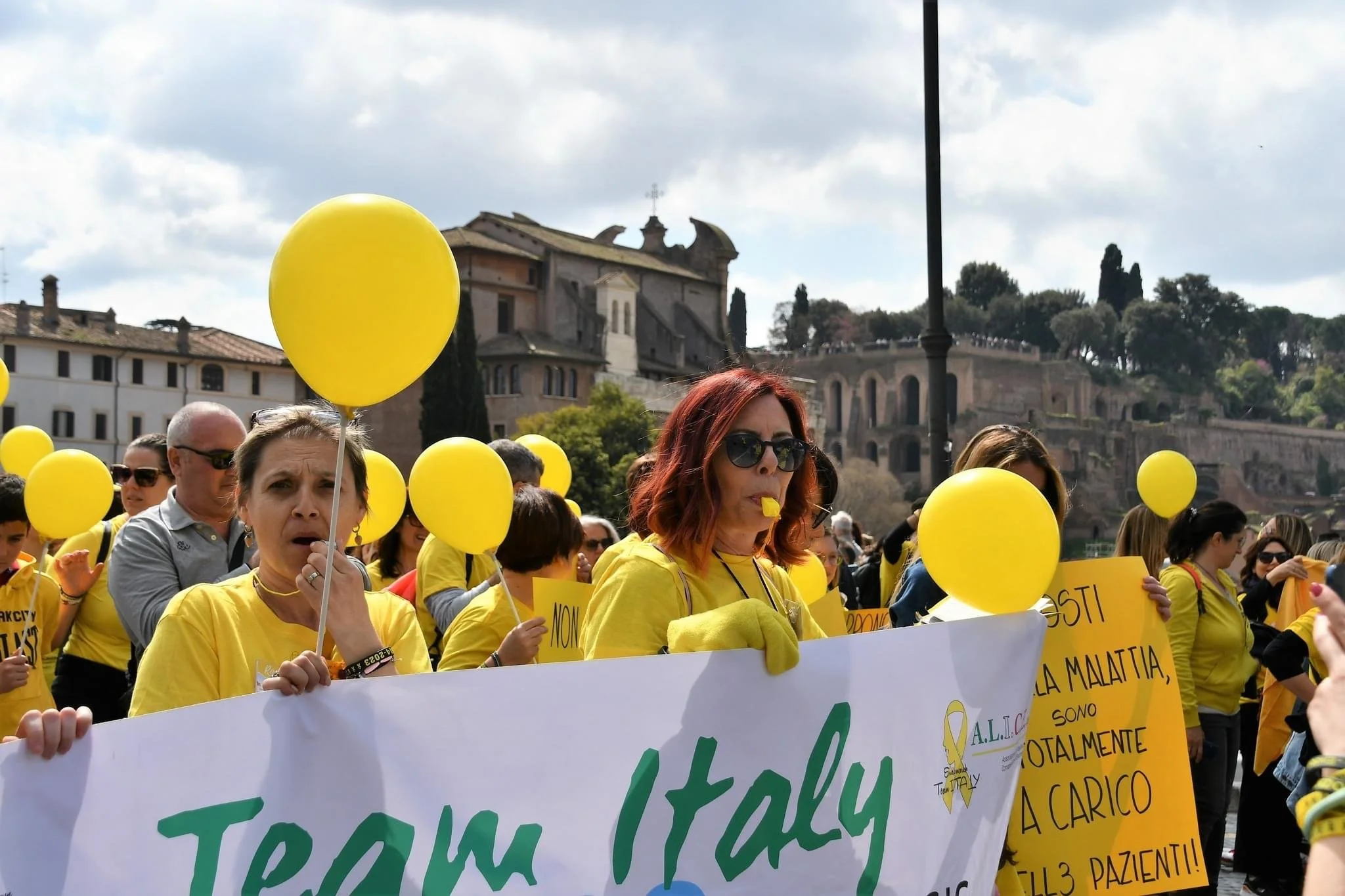 Gruppo di persone che partecipano a una manifestazione, alcune tengono palloncini gialli e cartelli, indossano magliette gialle, con uno sfondo di edifici storici.
