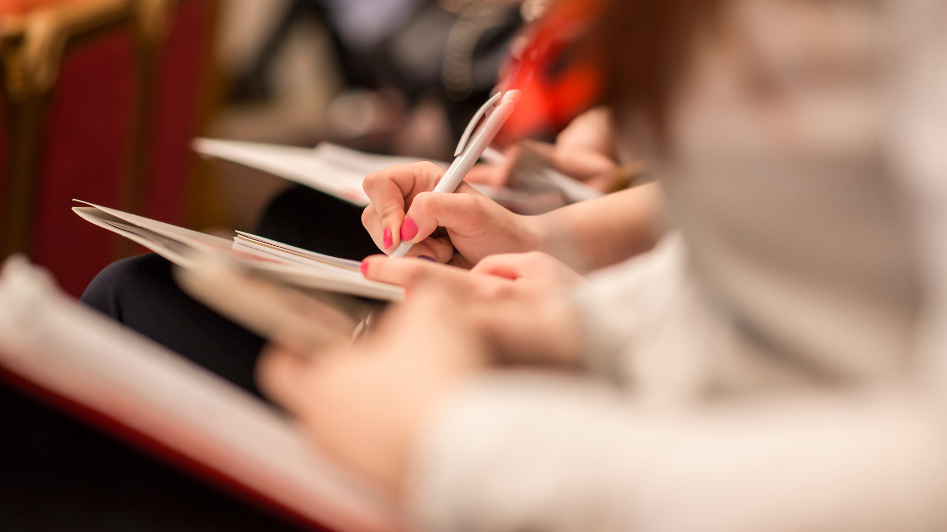 Persone che scrivono su quaderno con penna in un ambiente di conferenza o riunione.