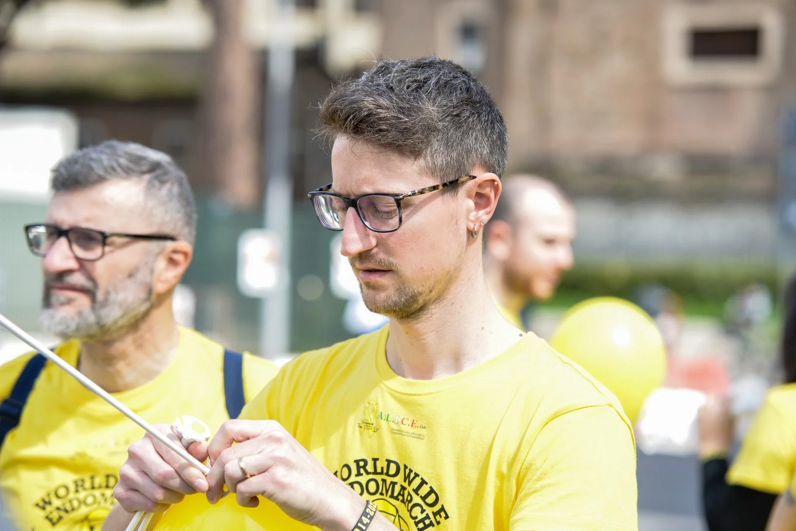 Un giovane uomo con occhiali e orecchini indossa una maglietta gialla durante un evento all'aperto. È concentrato mentre tiene in mano un oggetto, con altri individui in sfondo, anche loro in maglietta gialla.