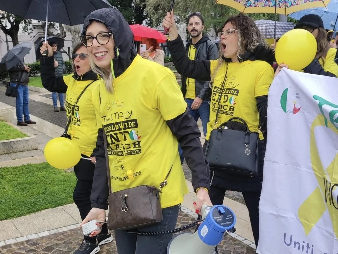 Un gruppo di persone partecipa a una manifestazione sotto la pioggia, indossando magliette gialle con scritte e portando palloncini e ombrelli.