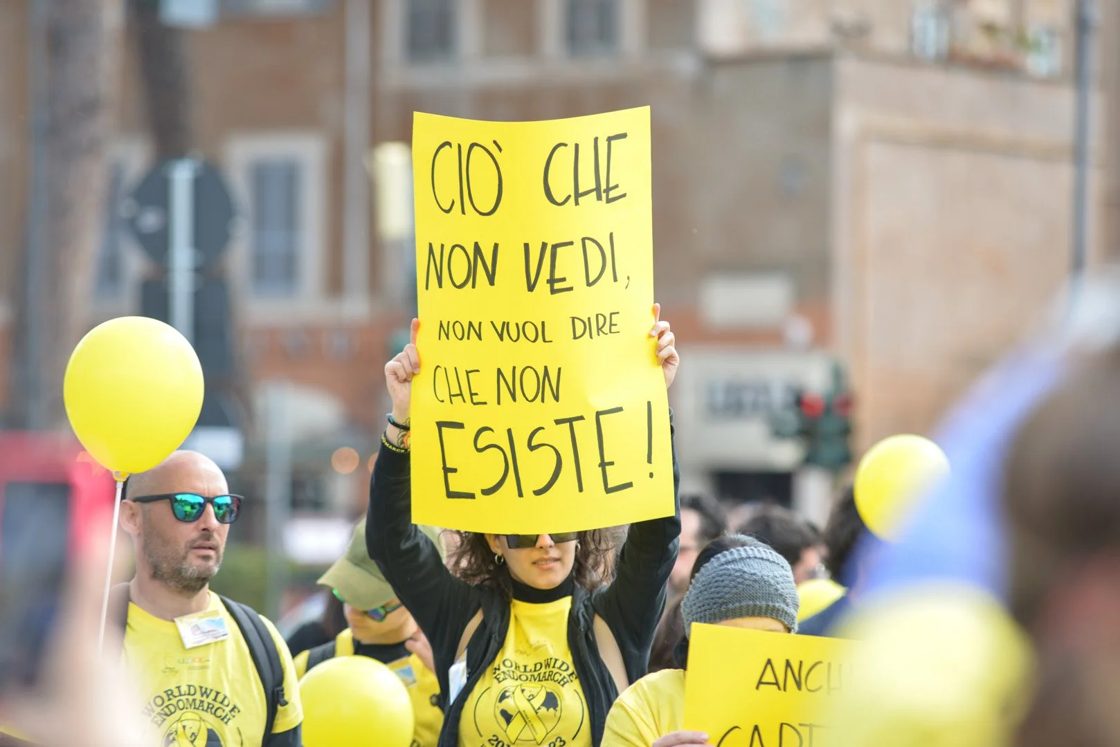 Una donna tiene un cartello giallo con la scritta 'CIO CHE NON VEDI, NON VUOL DIRE CHE NON ESISTE!', durante una manifestazione con molte persone che indossano magliette e palloncini gialli.