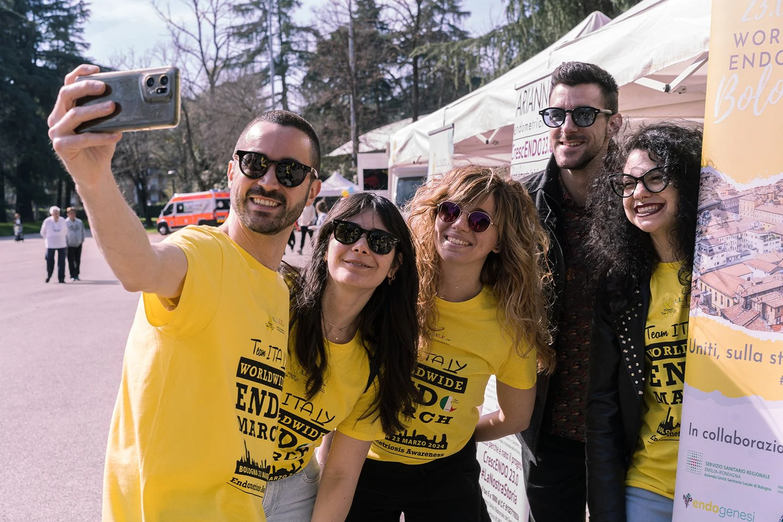 Gruppo di persone sorridenti che fanno un selfie all'aperto durante un evento, indossando magliette gialle con scritte riguardanti la consapevolezza dell'endometriosi, con alcuni in piedi accanto a uno stand con tenda.