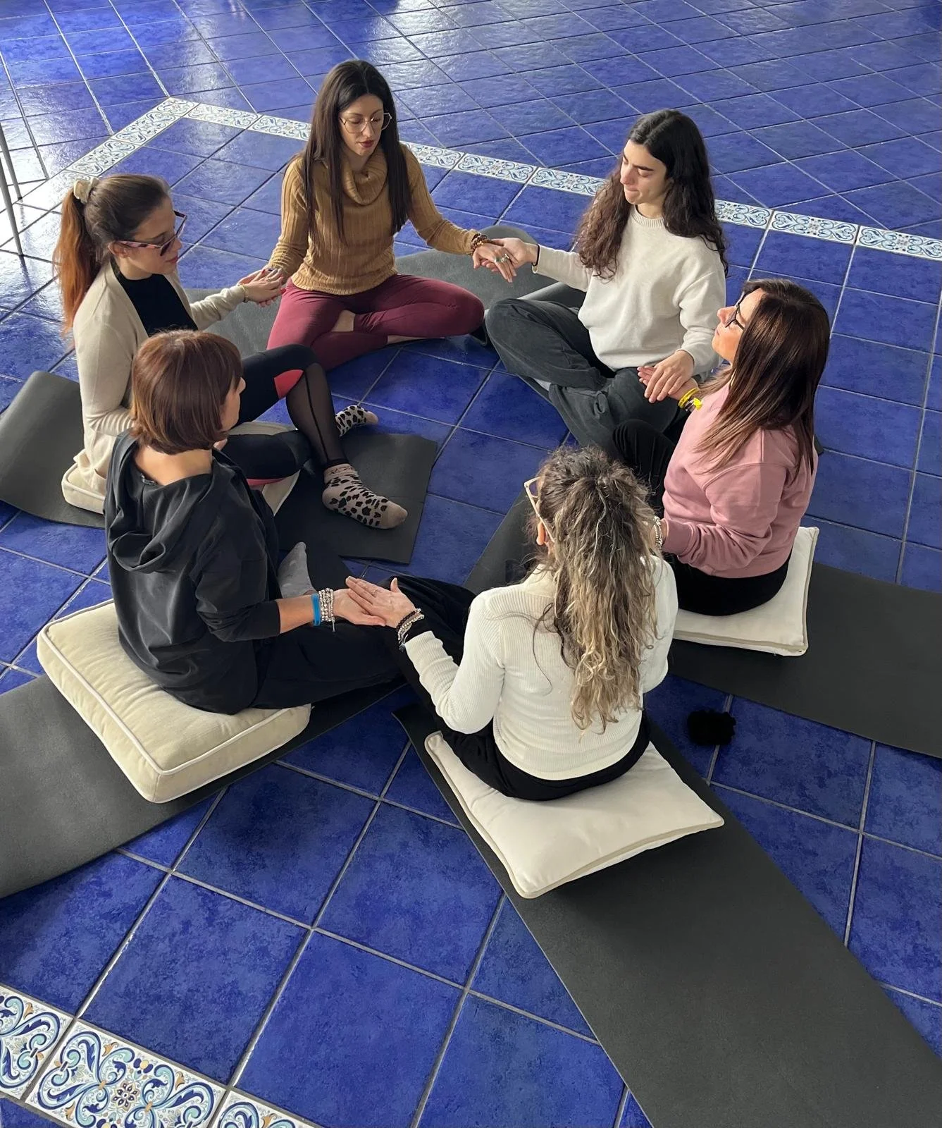 Sette donne sedute a cerchio su tappetini blu, che tengono le mani in un gesto di supporto e connessione