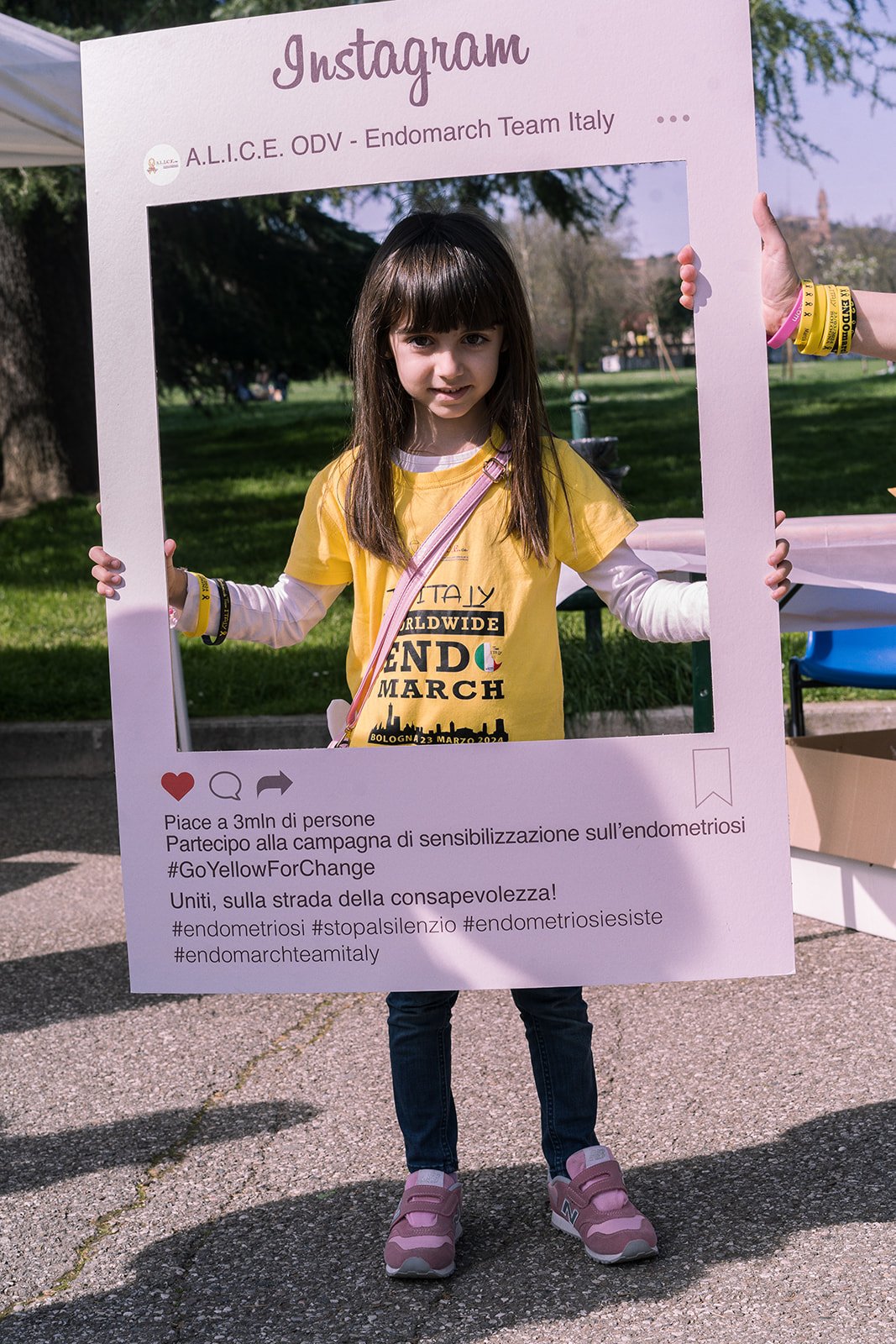 Bambina con maglietta gialla e pantaloni blu che tiene un cartellone gigante con scritte in italiano, durante un evento di sensibilizzazione, con un parco sullo sfondo.