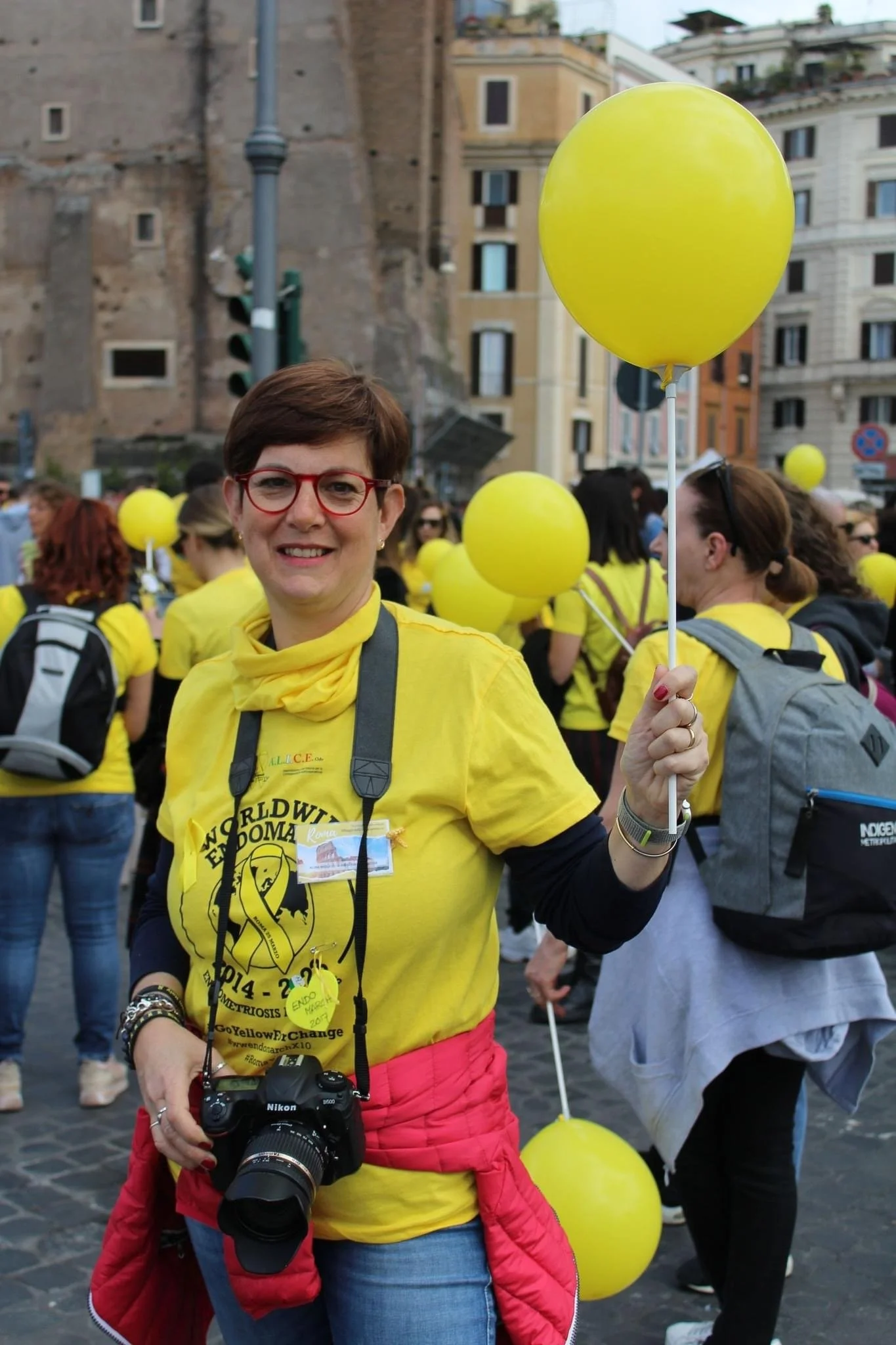 Una donna sorridente con occhiali rossi indossa una maglietta gialla con scritte e ha una fotocamera pendente al collo, durante una manifestazione o evento di sensibilizzazione. È in mezzo a un gruppo di persone vestite di giallo che tengono pallonci