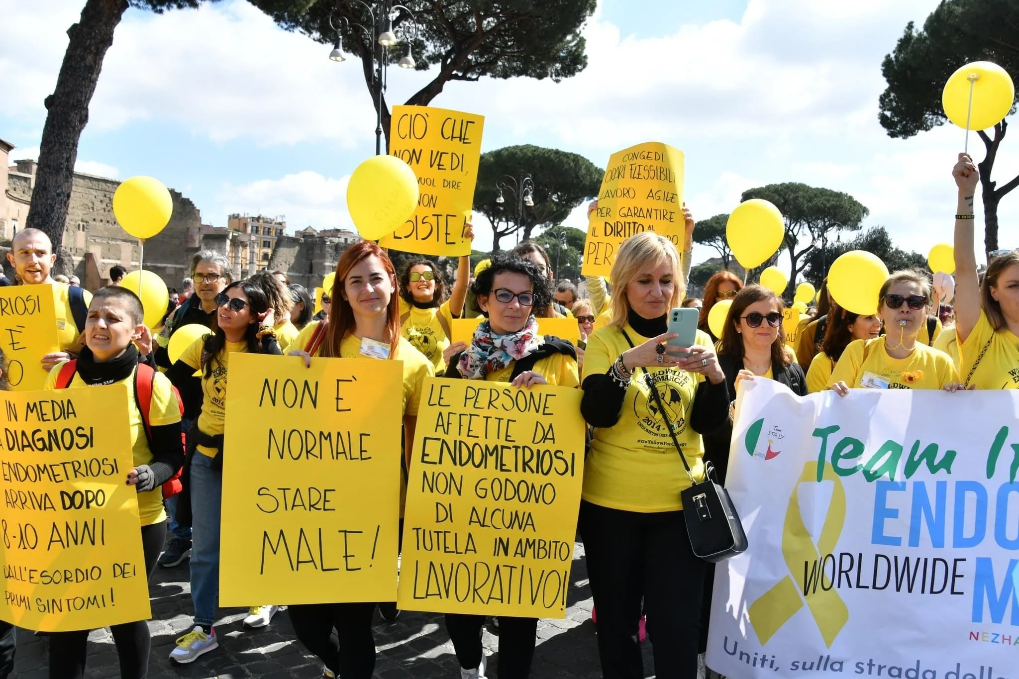 Gruppo di manifestanti con cartelli e palloncini gialli che protesta per i diritti delle donne e la consapevolezza sulla endometriosi, in un'area urbana con alberi e edifici sullo sfondo.