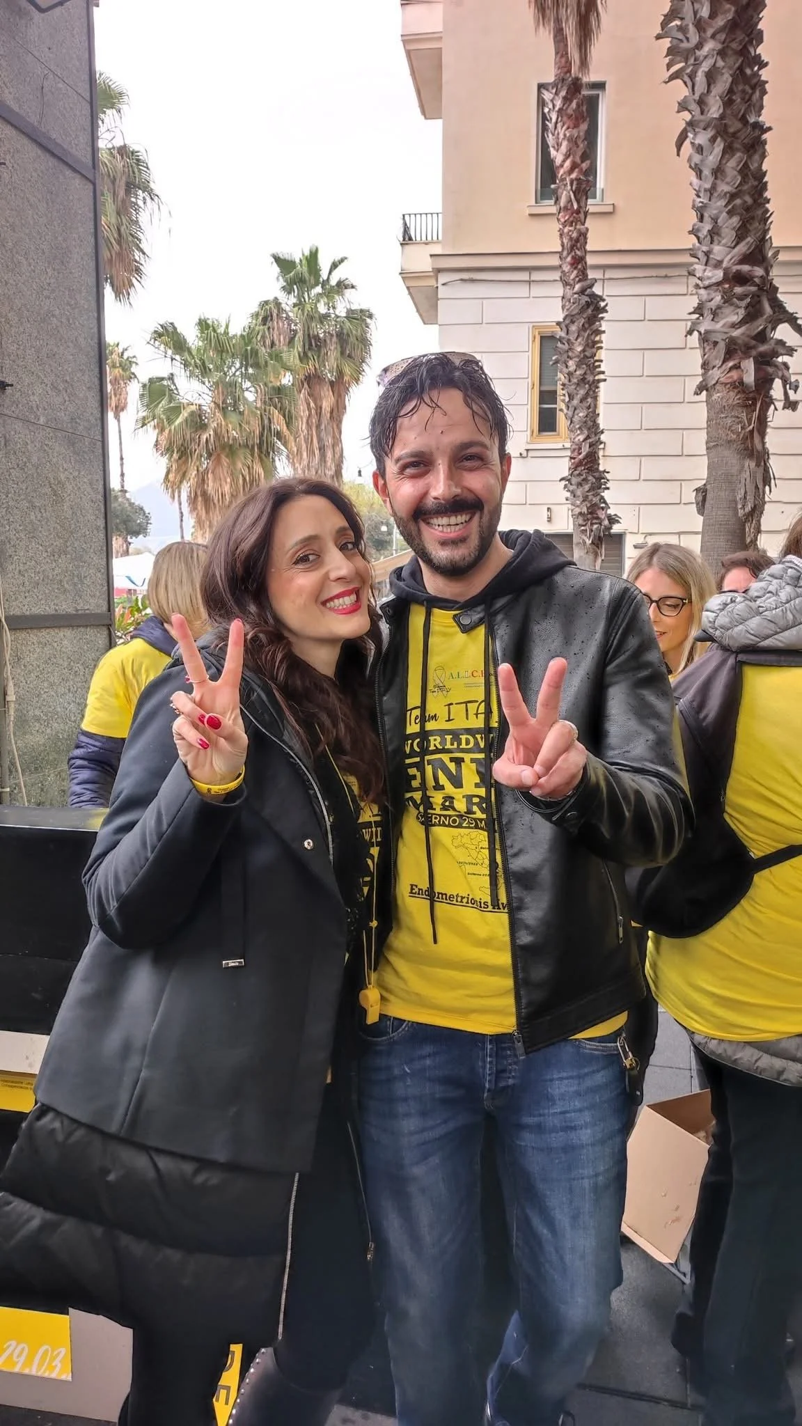 Due persone sorridenti e che fanno il gesto della pace, una donna con capelli ricci e una giacca e un uomo con barba e capelli corti vestito con una giacca di pelle e una maglietta gialla, in un contesto all'aperto con palme sullo sfondo.