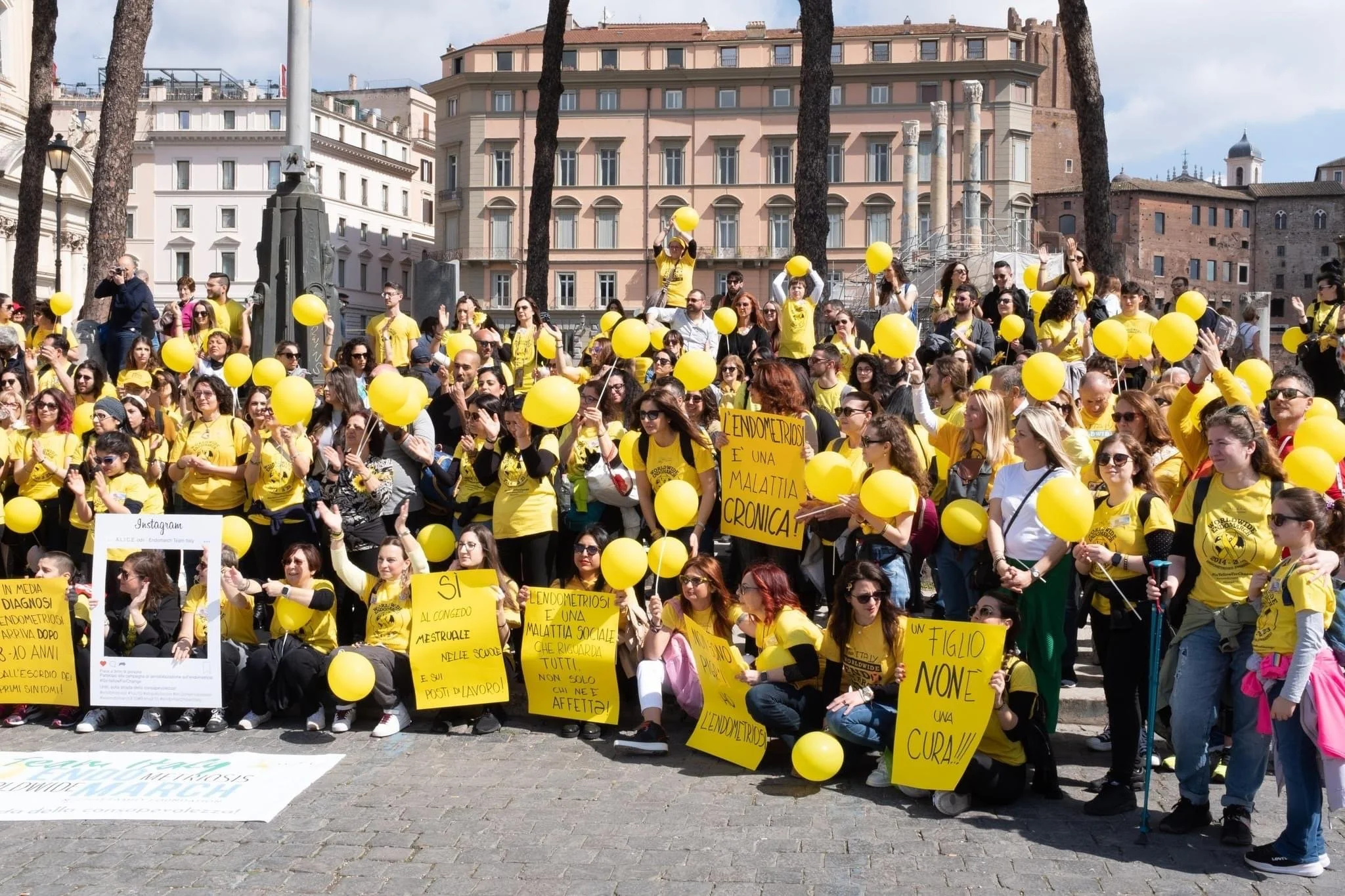 Gruppo di persone radunate in piazza, indossano magliette gialle e tengono palloncini gialli, alcune tengono cartelli con messaggi in italiano sulla endometriosi