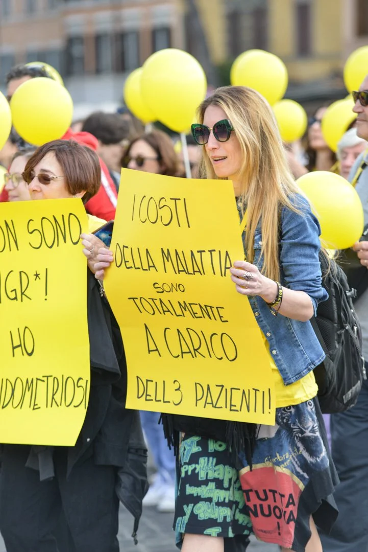 Gruppo di persone partecipanti a una manifestazione, molte con palloncini gialli, che tengono cartelli con messaggi di protesta.