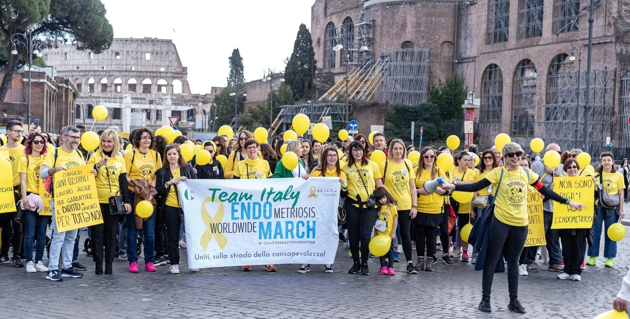 Un gruppo di persone partecipa a una marcia in Roma, con molti indossano magliette gialle e tengono palloncini gialli, e alcuni tengono cartelli con messaggi a favore della consapevolezza sull'endometriosi.