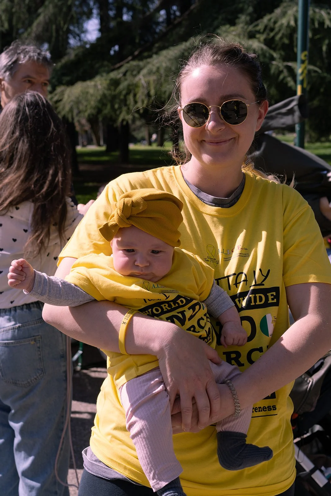 Una donna con occhiali da sole sorride mentre tiene in braccio un bambino vestito di giallo, entrambi in un parco durante una giornata di sole.