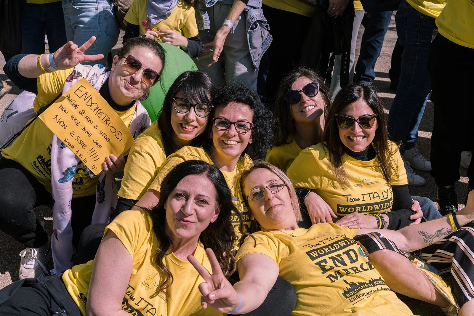 Gruppo di donne sorridenti indossando magliette gialle durante un evento all'aperto, alcune con occhiali da sole, alcune che fanno segno di pace, alcune con cartelli e braccialetti, sembra una manifestazione o una festa.
