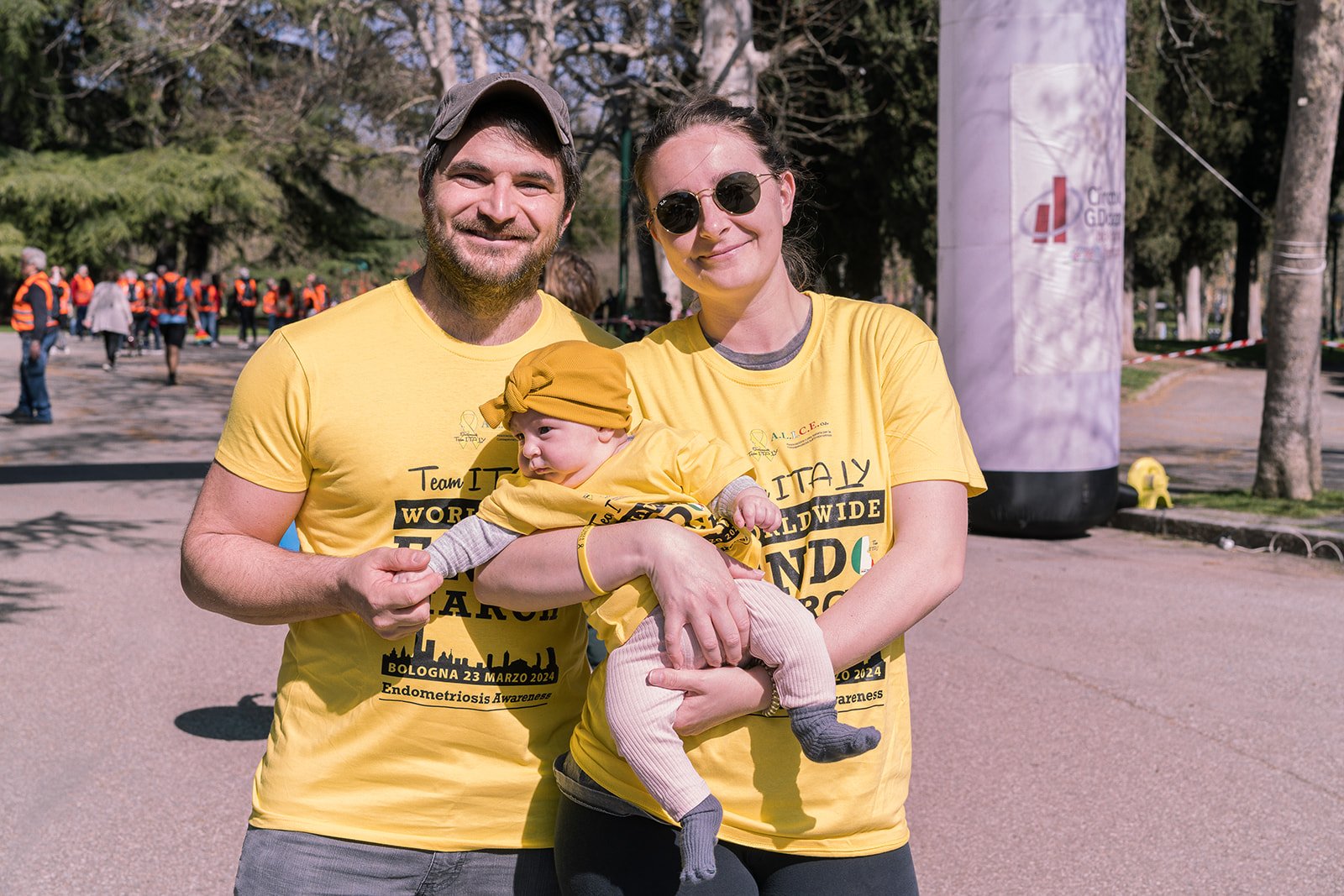 Una famiglia felice con due adulti e un bambino, tutti indossano magliette gialle durante una corsa benefica a Bologna per la consapevolezza dell'endometriosi, con molte persone sullo sfondo in un parco.