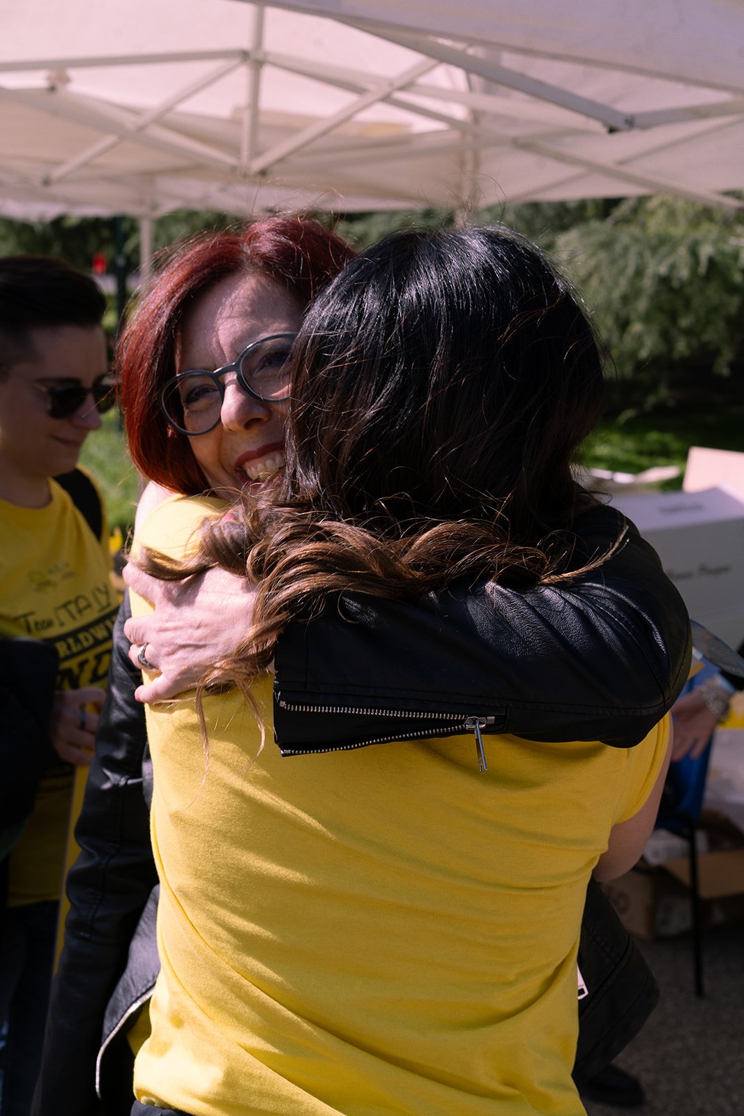 Due donne si abbracciano sorridendo sotto una tettoia bianca, una indossa una maglietta gialla e occhiali, l'altra una giacca in pelle nera.