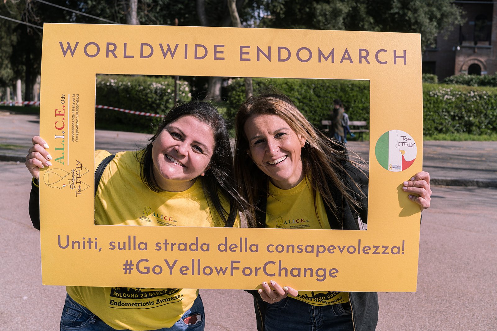 Due donne sorridenti tengono un grande frame giallo con scritto 'Worldwide Endomarch' e altre frasi, in occasione di una manifestazione di sensibilizzazione sulla endometriosi, indossando magliette gialle con scritte.