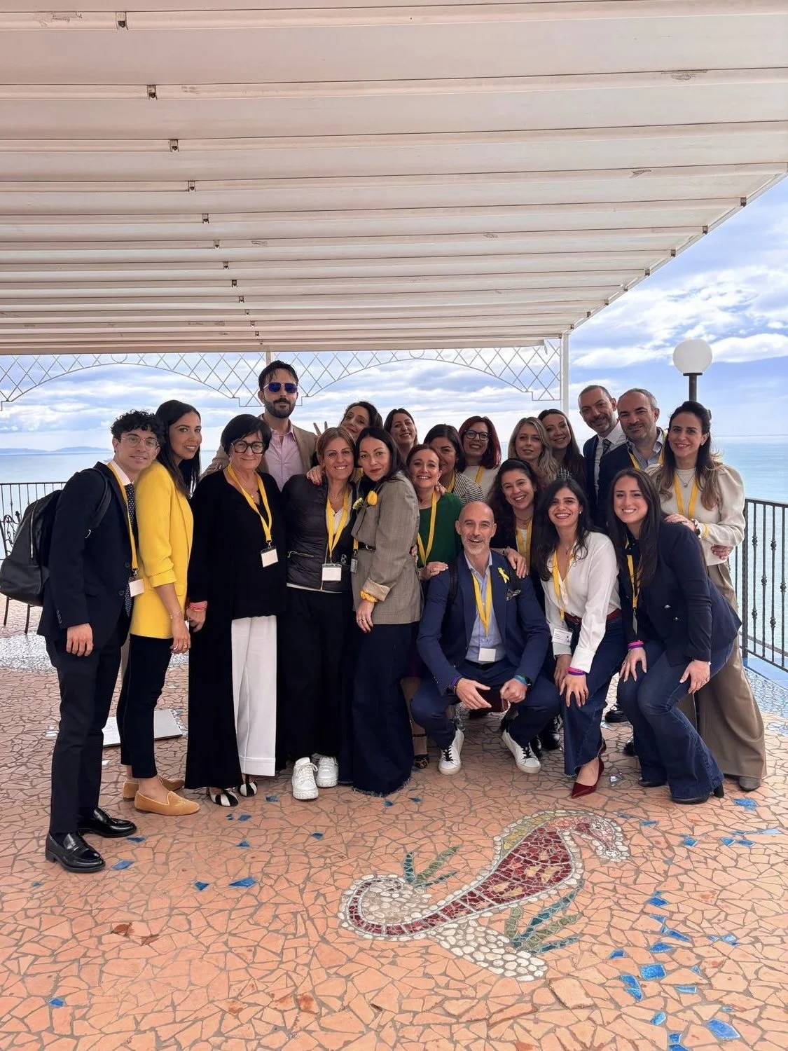 Gruppo di persone sorridenti in posa di fronte a un panorama di mare, alcune indossano magliette e sciarpe gialle, sotto un pergolato bianco.