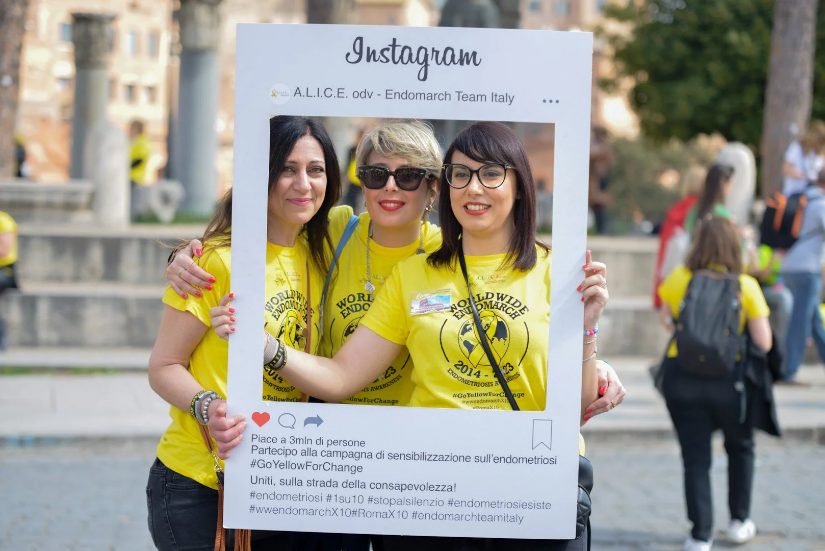 Tre donne che partecipano a una campagna di sensibilizzazione sull'endometriosi, indossano magliette gialle e tengono un cartellone a forma di cornice di un post di Instagram durante un evento all'aperto.