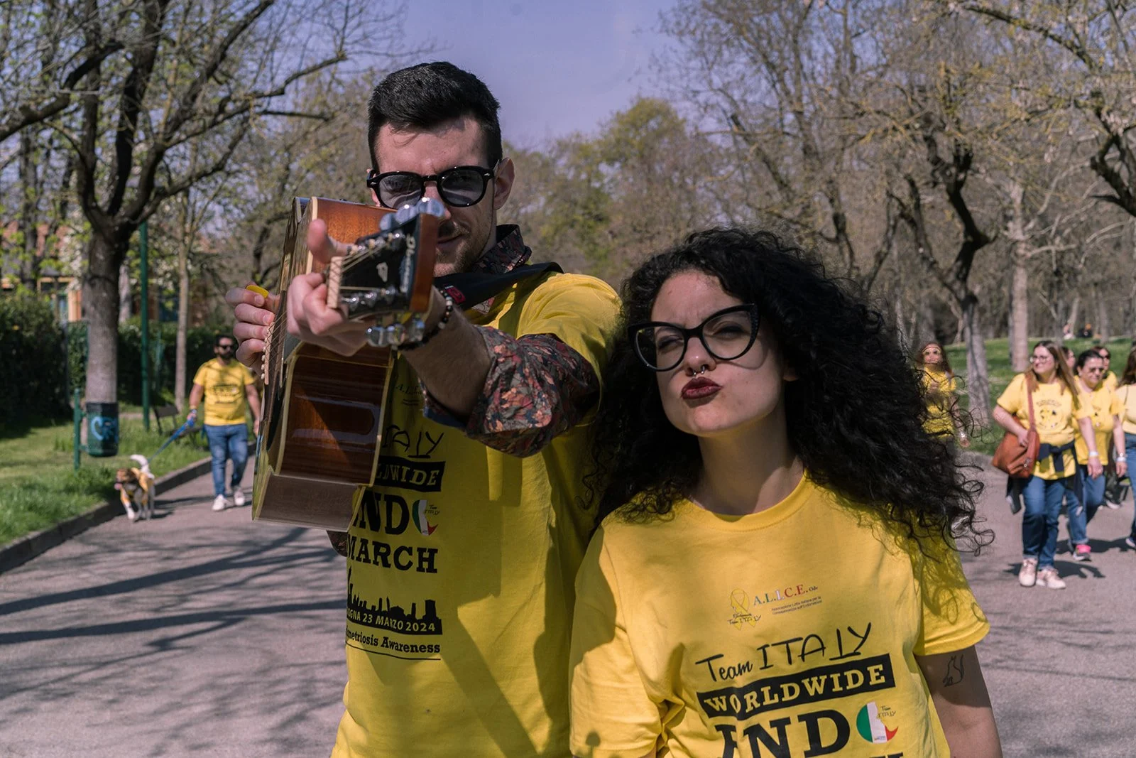 Due persone in primo piano, uno con una chitarra e occhiali da sole, e l'altra con occhiali neri e labbra che fanno un'espressione divertente. Entrambi indossano magliette gialle con scritte sulla partecipazione a una marcia mondiale. Sullo sfondo ci