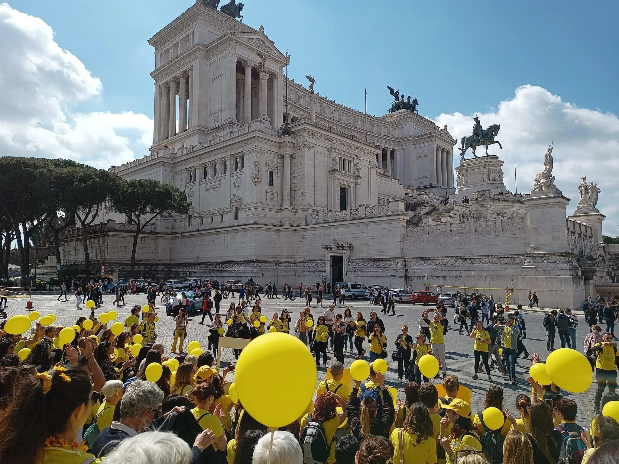 Gruppo di persone con palloncini gialli davanti al Altare della Patria, un grande monumento bianco con statue e colonne a Roma.