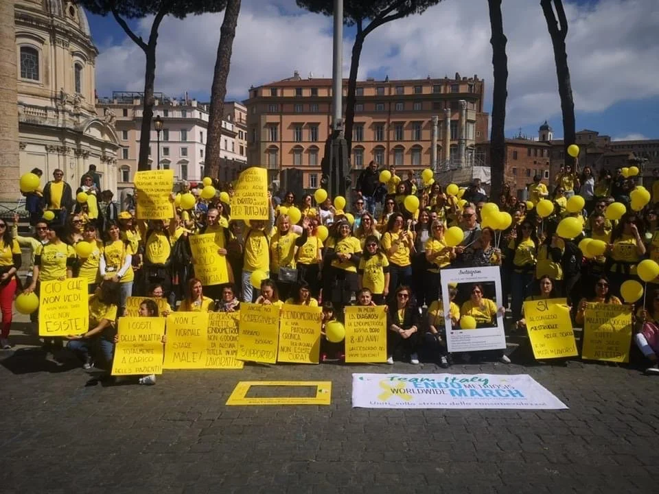 Gruppo di persone che partecipano a una manifestazione in piazza, molte vestite con magliette gialle e che tengono palloncini gialli. Presentano cartelli e un grande striscione con scritto "Team Pedia" e "Worldwide March".
