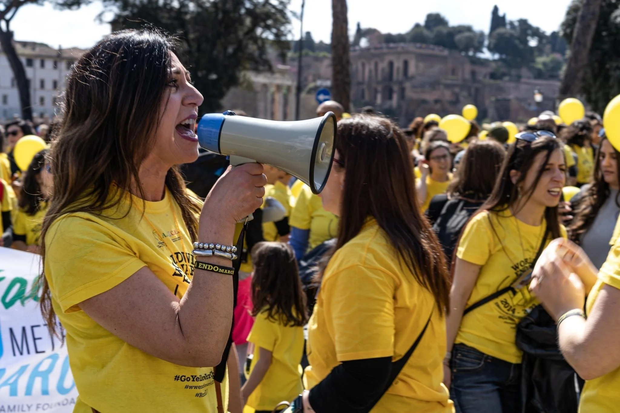 Donna che parla in un megafono durante una manifestazione con molte persone vestite di giallo e palloncini gialli.