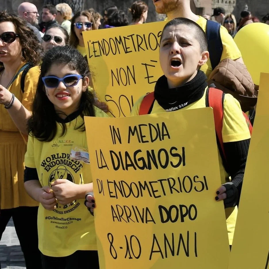 Ragazza con occhiali da sole e sorriso, ragazzo con espressione arrabbiata che tiene un cartello giallo durante una manifestazione di sensibilizzazione sull'endometriosi, altri partecipanti con cartelli e palloncini gialli sullo sfondo.