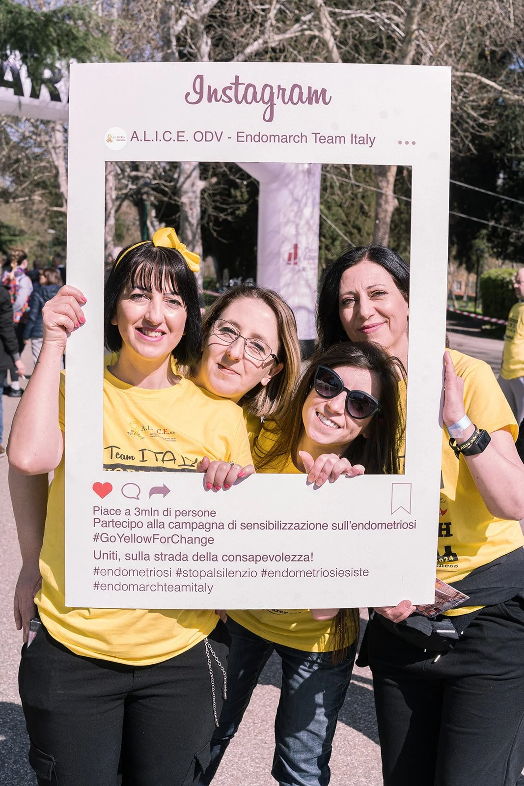 Gruppo di donne sorridenti che tengono un grande fotogramma a forma di post di Instagram, nel contesto di una manifestazione all'aperto sulla sensibilizzazione sull'endometriosi, con alberi e altre persone sullo sfondo.