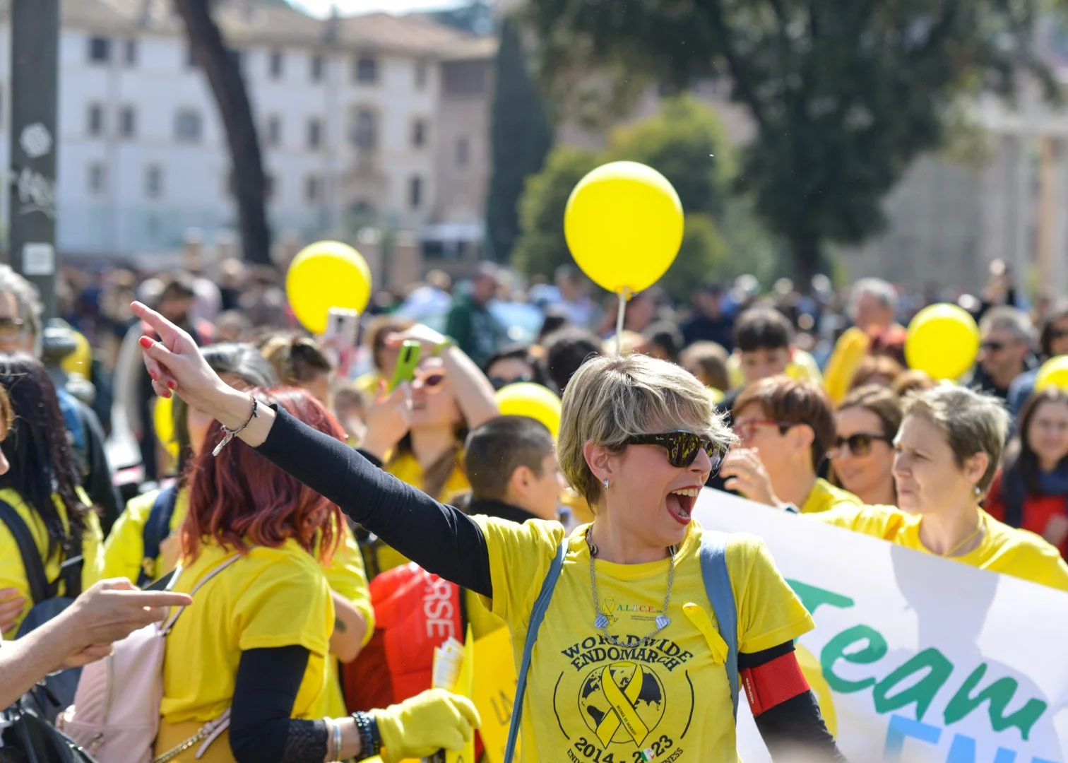 Gruppo di persone che partecipano a una marcia o maratona, molti indossano magliette gialle con scritte e portano palloncini gialli all'aperto in una giornata di sole.
