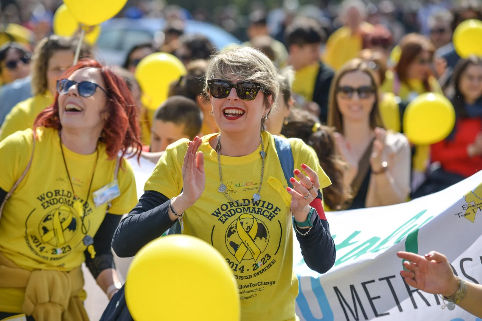 Diverse persone partecipano a una marcia benefica, indossando t-shirt gialle con logo e slogan, e tengono palloncini gialli durante un evento di sensibilizzazione.