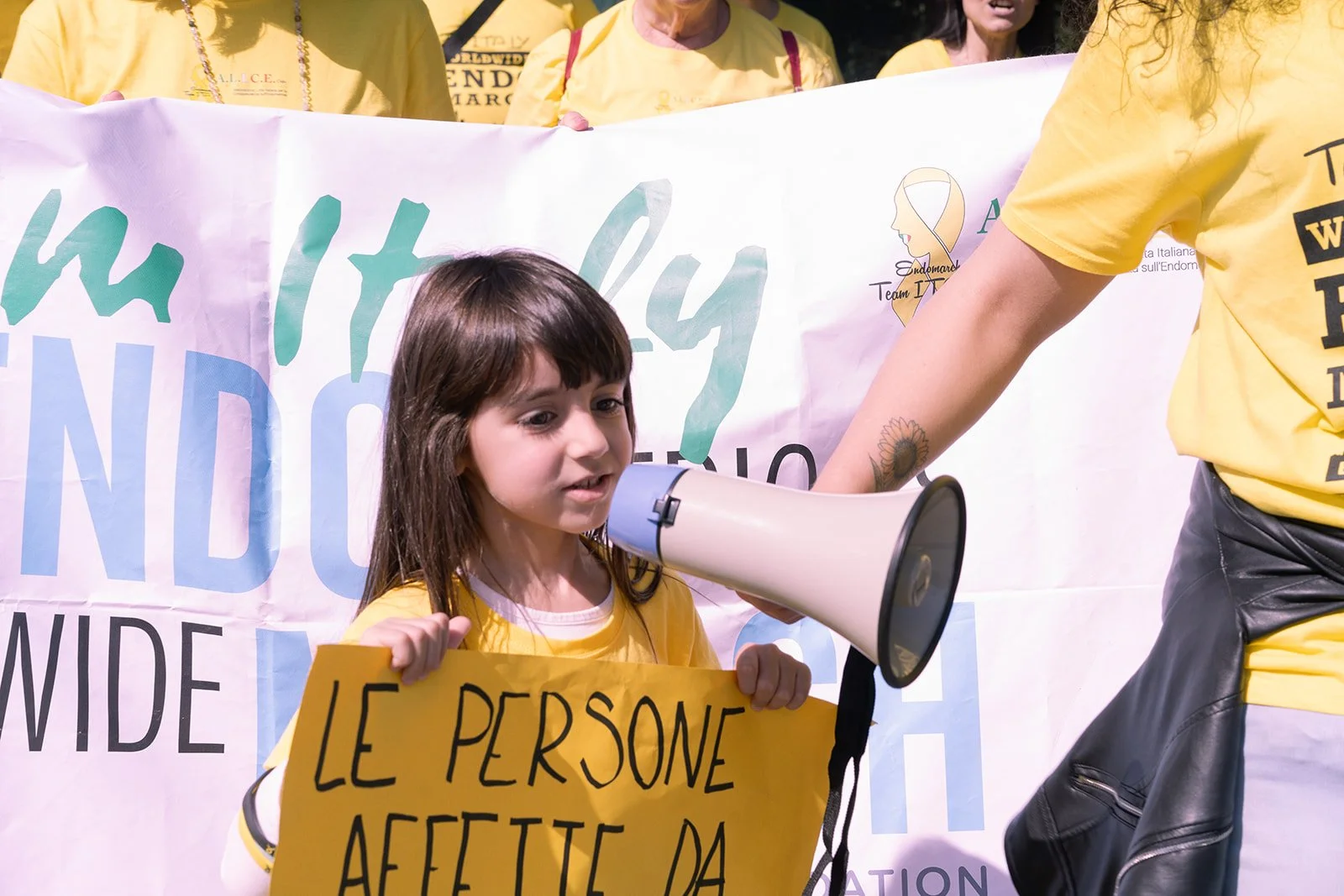 Una bambina tiene in mano un cartello giallo con scritto 'LE PERSONE AFFETTE DA'. Sta parlando attraverso un megafono portato da un'altra persona, partecipando a una manifestazione di sensibilizzazione.