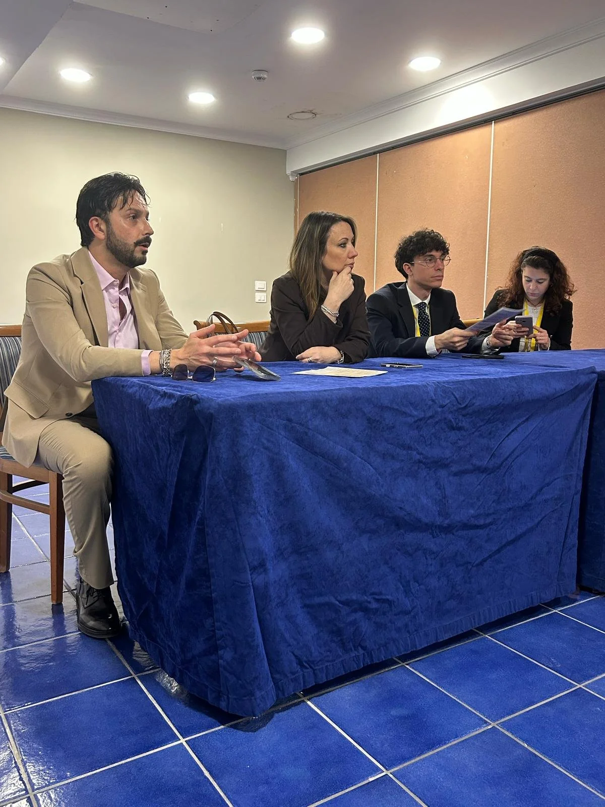 Quattro persone sedute dietro un tavolo coperto da una tovaglia blu in una sala conference, una donna e tre uomini durante una riunione o conferenza.